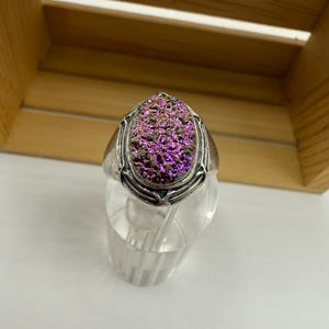 May include: A silver ring with an oval-shaped, purple and brown druzy stone. The ring has an ornate design around the stone. The ring is displayed on a clear, cylindrical stand against a wooden background.