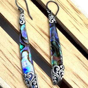 May include: A pair of long, dangling earrings with iridescent abalone shells. The earrings have a silver-colored metal setting with decorative scrollwork at the top and bottom. The abalone shells display a mix of blue, green, and purple hues.