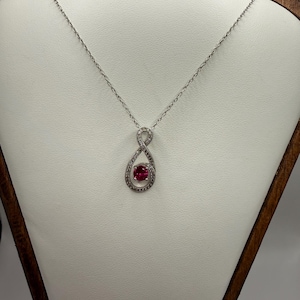 May include: A silver-toned necklace featuring a delicate chain and a pendant shaped like a twisted infinity loop. The pendant is encrusted with small clear stones and centers a single, round, vibrant pink gemstone at the bottom of the loop.