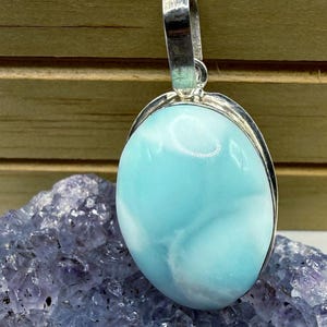 May include: An oval-shaped, polished light blue Larimar gemstone pendant set in a smooth silver metal frame. The stone features swirling white patterns throughout its surface and includes a silver metal bail at the top for hanging.