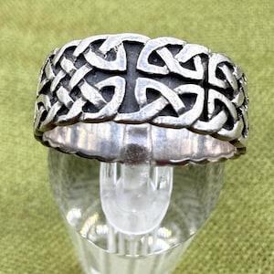 May include: A silver-toned ring with a Celtic knot design. The ring features a repeating pattern of interwoven knots and crosses. The ring is displayed on a clear, glass stand against a green background.