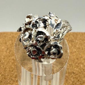 May include: A silver ring featuring a cluster of detailed floral elements. The ring's design includes multiple flower shapes and leaf accents, creating a textured, nature-inspired aesthetic. The ring is displayed on a clear stand.