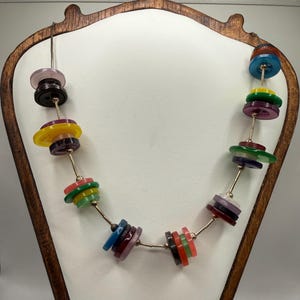 May include: A necklace featuring a series of stacked, multi-colored plastic buttons in shades of red, orange, yellow, green, blue, purple, and pink, strung together on a thin metallic wire with small gold-toned spacer beads between each group.