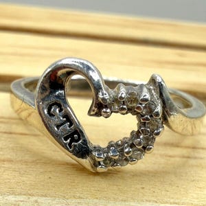 May include: A silver-toned ring featuring a heart-shaped design. The left side of the heart is smooth and engraved with the letters CTR, while the right side is encrusted with a series of small, clear, round-cut stones set in a metallic band.
