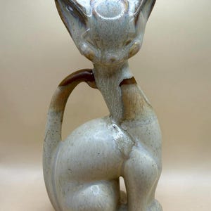 May include: A ceramic cat figurine in a seated pose. The cat has a cream-colored body with brown and blue accents. The ears are pointed, and the tail curves around the body. A decorative item.