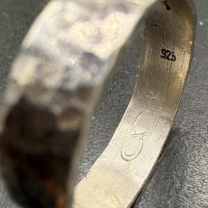 May include: A silver ring with a hammered, textured exterior surface. The interior of the band features a stamped 925 hallmark indicating sterling silver composition and an engraved logo consisting of two interlocking circular shapes.