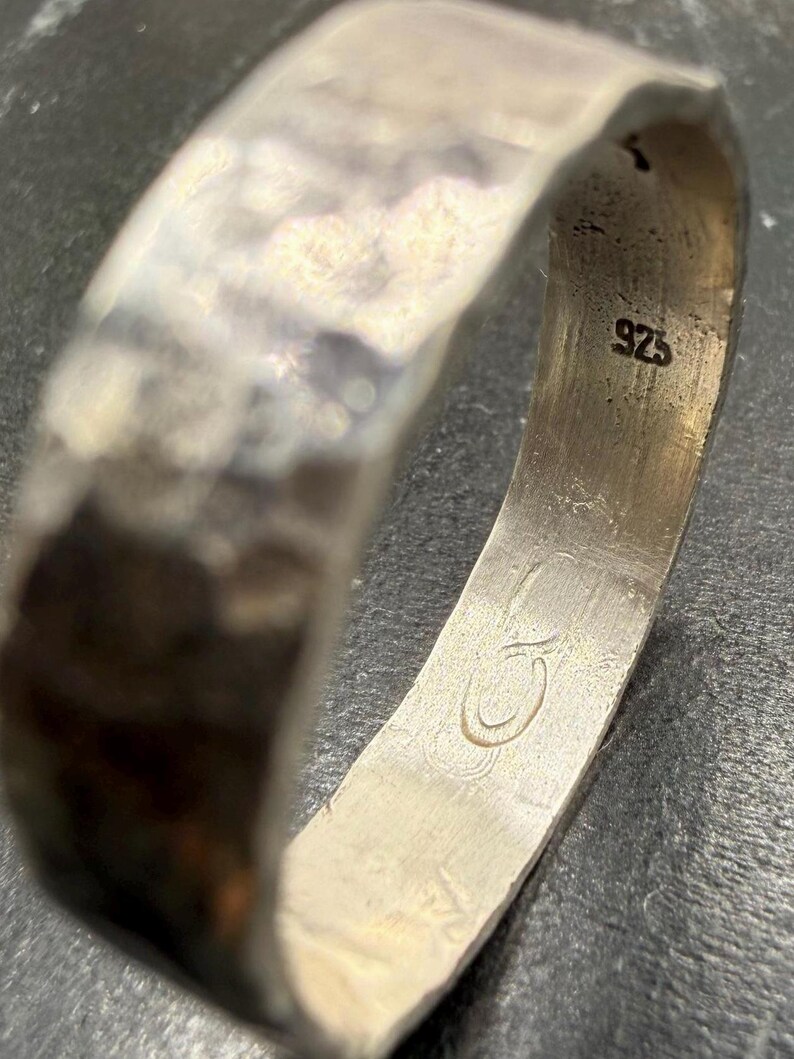 May include: A silver ring with a hammered, textured exterior surface. The interior of the band features a stamped 925 hallmark indicating sterling silver composition and an engraved logo consisting of two interlocking circular shapes.