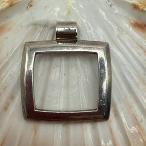 May include: A square silver pendant with a hollow center and a top bail for attachment. The metal surface is smooth and reflective, featuring a slightly curved frame design that creates a geometric opening in the middle of the piece.