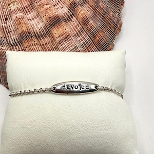 May include: A silver bracelet with a beaded chain and an oval charm. The charm is engraved with the word "devoted" in a simple, elegant font. The bracelet is displayed on a white surface, with a seashell in the background.