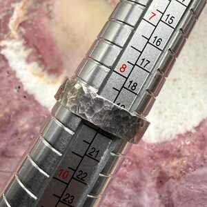 May include: A silver-toned ring with a hammered, textured surface placed on a metal ring mandrel. The mandrel features clear measurement markings, showing the ring rests between size 8 and 9 on the standard American ring sizing scale.