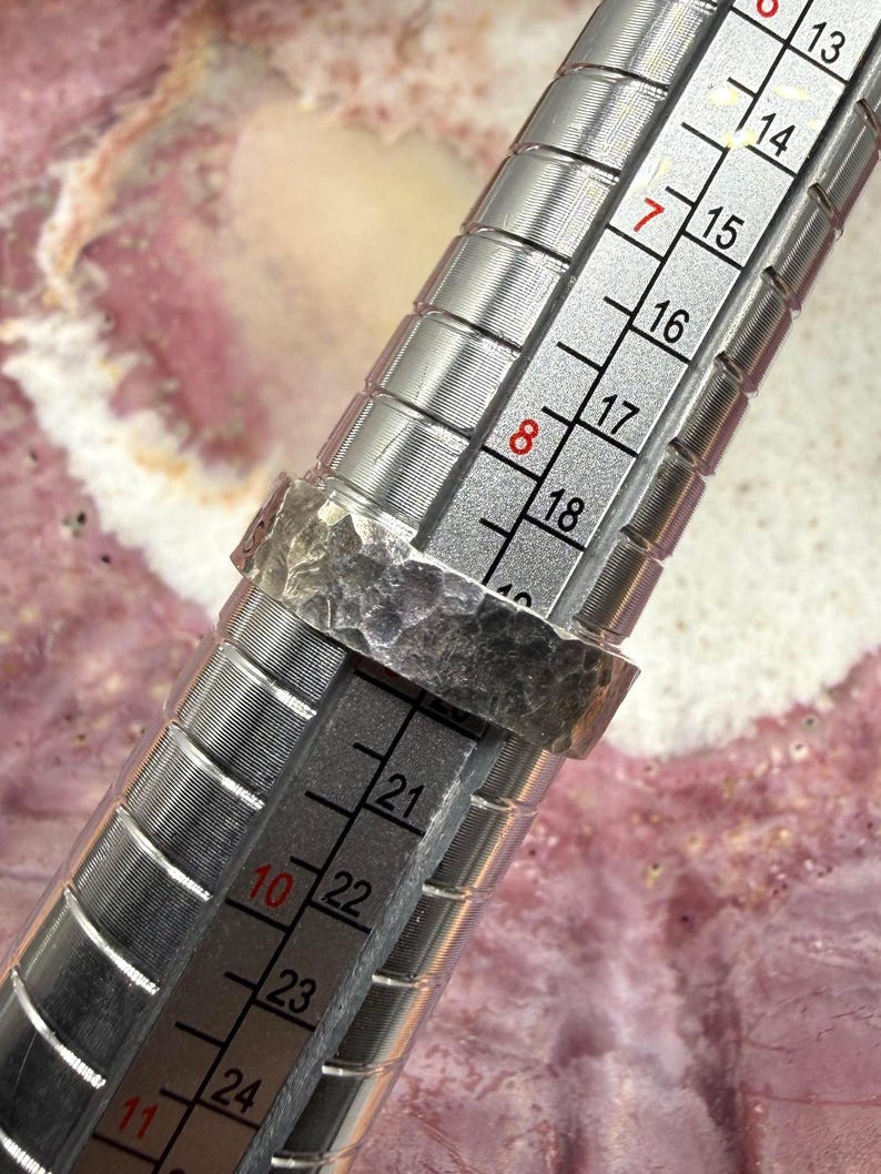 May include: A silver-toned ring with a hammered, textured surface placed on a metal ring mandrel. The mandrel features clear measurement markings, showing the ring rests between size 8 and 9 on the standard American ring sizing scale.