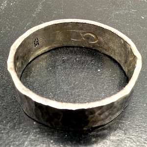 May include: A silver ring with a hammered, textured exterior surface. The interior of the band features a stamped 925 hallmark indicating sterling silver composition and an additional engraved symbol resembling a stylized infinity loop or link.