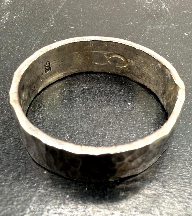 May include: A silver ring with a hammered, textured exterior surface. The interior of the band features a stamped 925 hallmark indicating sterling silver composition and an additional engraved symbol resembling a stylized infinity loop or link.
