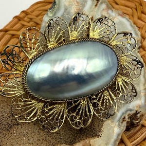 May include: An oval brooch featuring a smooth, iridescent gray cabochon center surrounded by a gold-toned metal filigree frame with a scalloped petal design. The piece has a twisted rope-style border encircling the central stone.