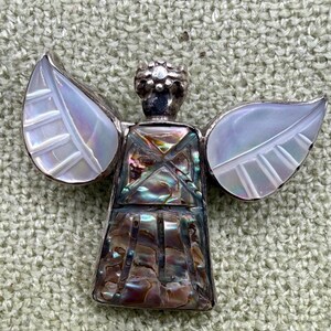 May include: A silver-toned angel pin with iridescent abalone shell accents. The angel has detailed wings and a crown. The body of the angel is made of abalone shell, with a silver border. The wings are white with leaf-like details.