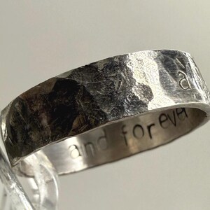 May include: A silver-toned metal ring with a heavily hammered, textured exterior surface. The interior band features the engraved text "and forever" in a simple, lowercase sans-serif font. The ring has a wide, flat band profile.