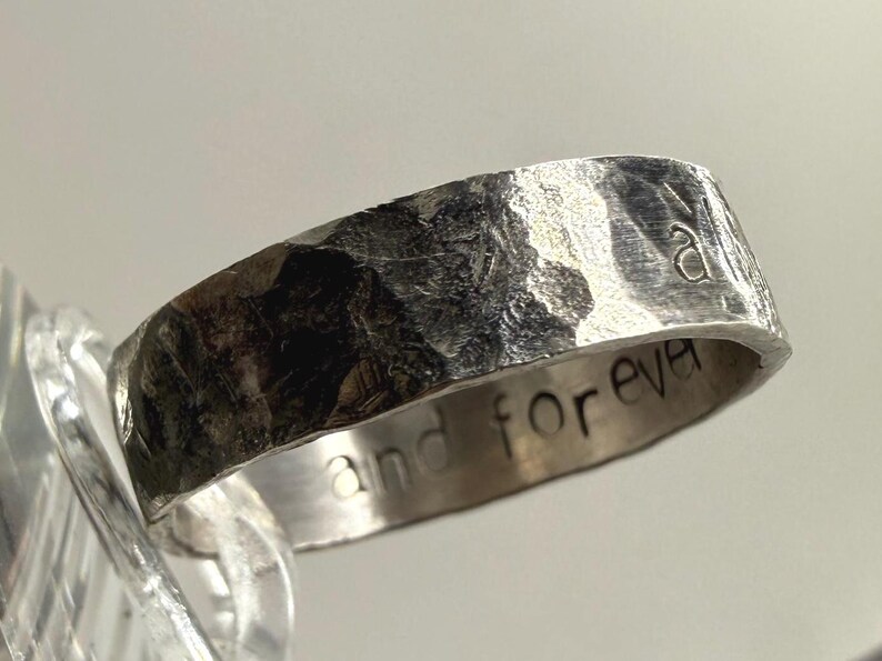 May include: A silver-toned metal ring with a heavily hammered, textured exterior surface. The interior band features the engraved text "and forever" in a simple, lowercase sans-serif font. The ring has a wide, flat band profile.
