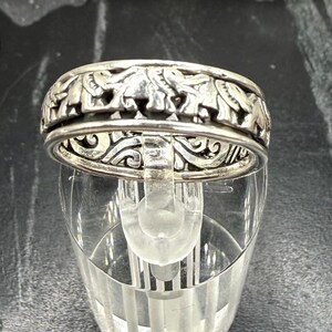 May include: A silver-toned metal ring featuring a repeating embossed pattern of elephants around the outer band. The ring has a secondary inner band layer visible through the cutouts, displaying an intricate, swirling, scroll-like engraved design.