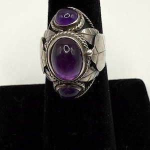 May include: A silver ring featuring three oval, deep purple amethyst stones. The ring has a detailed design with leaf-shaped accents and a textured band. The gemstones are set in a decorative setting.