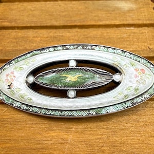 May include: Oval-shaped brooch with a detailed design. The brooch features a central green and gold design, surrounded by a white and silver border with floral accents. The brooch is set against a wooden background.