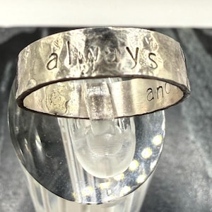 May include: A silver-toned metal ring with a hammered, textured surface. The word "always" is stamped in lowercase letters across the front outer band, while the word "and" is partially visible on the right side of the inner band surface.