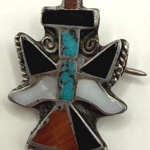 May include: A silver-toned pin with a cross-like design. The pin features inlaid sections of black, white, turquoise, and orange stones. The edges are detailed with a twisted rope design.