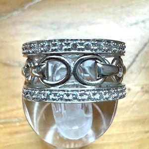 May include: A silver ring with a decorative design featuring a series of interlocking links. The ring is encrusted with small, clear gemstones along the top and bottom edges, creating a sparkling effect. The ring is displayed on a clear, rounded object.