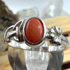 May include: A silver ring featuring an oval, reddish-orange coral stone. The ring has a decorative design on the band with a knot-like detail on the sides. The coral is set in a bezel setting, and the ring is displayed on a clear glass surface.