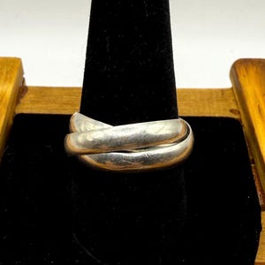 May include: A silver-toned ring composed of three interlocking bands that overlap each other. The metal surface has a smooth, polished finish with a reflective quality, creating a continuous, rounded shape that sits horizontally on the display.