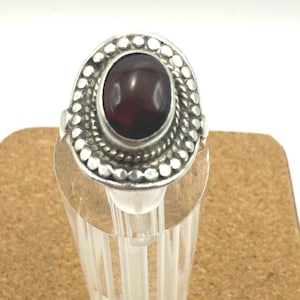 May include: A silver ring with an oval, dark red gemstone. The gemstone is surrounded by a beaded border and set in a silver band. The ring is displayed on a clear, cylindrical stand.