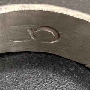 May include: A metallic ring featuring a brushed, matte silver-toned finish. The interior surface of the band displays a singular, engraved symbol that resembles a stylized, lowercase letter o or a small, circular loop etched into the metal.