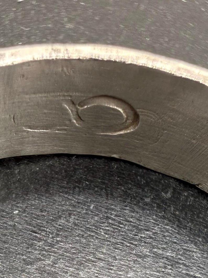 May include: A metallic ring featuring a brushed, matte silver-toned finish. The interior surface of the band displays a singular, engraved symbol that resembles a stylized, lowercase letter o or a small, circular loop etched into the metal.