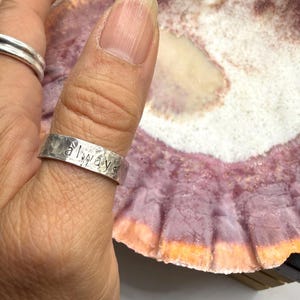 May include: A silver-colored metal ring with a hammered, textured finish and the word "always" stamped in lowercase letters on the outer band. The ring is worn on a thumb, positioned next to a large, textured seashell with purple and white hues.