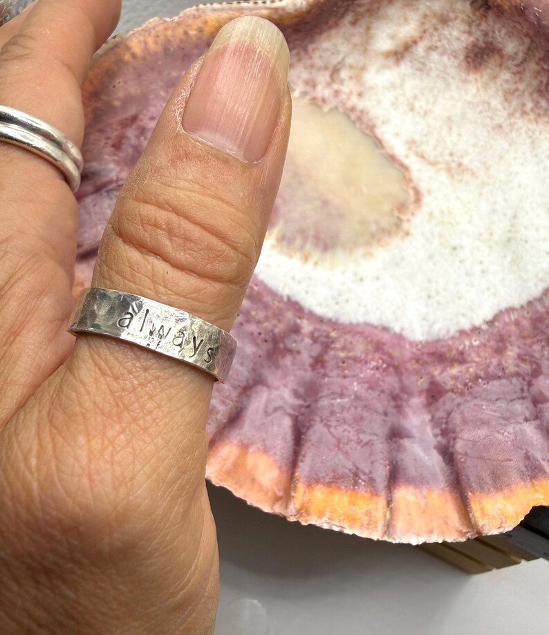 May include: A silver-colored metal ring with a hammered, textured finish and the word "always" stamped in lowercase letters on the outer band. The ring is worn on a thumb, positioned next to a large, textured seashell with purple and white hues.