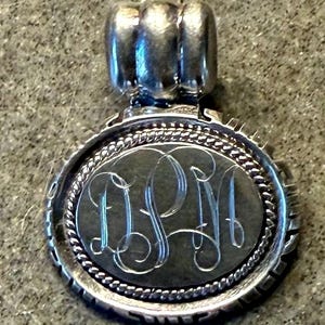 May include: A circular silver pendant featuring a central dark disc engraved with the cursive monogram letters DPM. The pendant is framed by a twisted rope border and a notched outer edge, topped with a smooth, rounded silver bail for hanging.