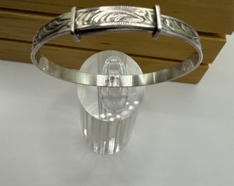 May include: A silver adjustable baby bangle bracelet. The bracelet features an engraved floral design on the central bar and decorative detailing along the sides. The bracelet is displayed on a clear acrylic stand against a wooden background.