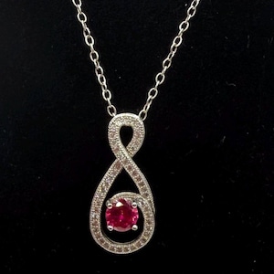 May include: A silver-toned pendant necklace featuring an infinity-shaped loop design encrusted with small clear stones. A single round, vibrant red gemstone is set within the lower curve of the pendant, which hangs from a delicate silver-toned chain.