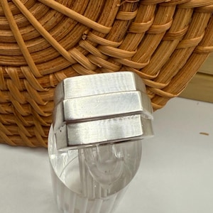 May include: A silver-colored ring with a rectangular design. The ring has a stacked appearance, with three horizontal bands. The ring is displayed on a clear plastic stand, with a woven basket in the background.