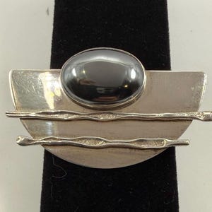 May include: A silver-toned brooch featuring a semi-circular base with a smooth, polished finish. The piece is accented by two horizontal, textured metal bars and a single, large, oval-shaped dark gray cabochon stone centered at the top.