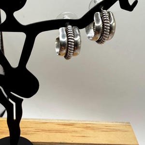 May include: A pair of silver hoop earrings featuring a smooth, wide outer band and a central, raised rope-textured detail. The earrings are finished with standard post-back closures, showcasing a polished metallic surface with subtle dark oxidation.