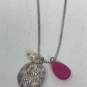 May include: A silver-toned ball chain necklace featuring three charms: a round, hammered metal disc engraved with the repeated word "HOPE," a small white spherical bead, and a smooth, teardrop-shaped pendant in a vibrant, translucent fuchsia pink.