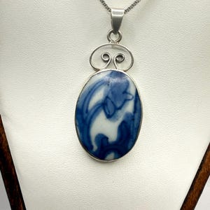 May include: A silver pendant necklace featuring an oval-shaped ceramic piece with a blue and white marbled pattern. The pendant is held by a silver bail with a decorative wire scroll design and hangs from a thin silver box chain necklace.
