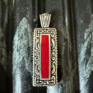May include: A rectangular silver pendant with a red center. The pendant has ornate detailing around the edges and a decorative bail at the top. The pendant is likely made of metal.