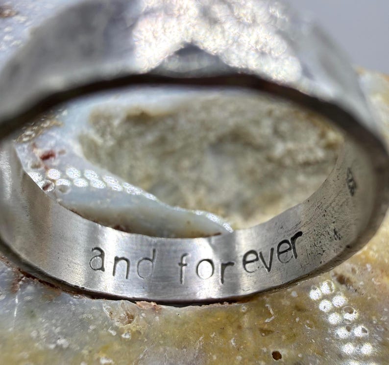 May include: A silver-toned metal ring with a hammered, textured exterior surface. The interior of the band features the engraved lowercase text "and forever" in a simple, sans-serif font. The ring has a wide, substantial band construction.