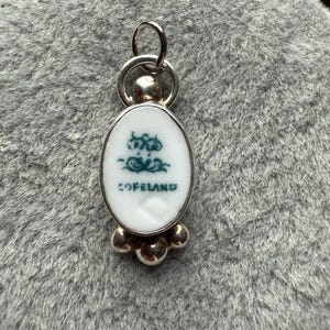 May include: A silver pendant with an oval, white ceramic insert featuring a green floral design and the word "COPELAND." The pendant has a loop at the top and a cluster of three silver beads at the bottom. The pendant is displayed on a gray, textured surface.