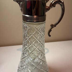 May include: A clear glass pitcher with a silver metal lid and handle. The pitcher has a diamond pattern design. The handle is ornate and curved. The pitcher is likely used for serving beverages.