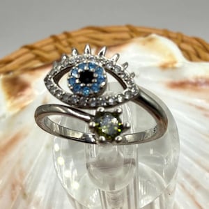 May include: A silver ring featuring an eye design with a blue iris and clear stone accents. The ring has a second green stone. The ring is displayed on a clear stand.