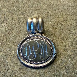 May include: Silver pendant with an oval shape and a decorative border. The pendant features an engraved monogram within the oval. The top of the pendant has a small, three-loop design for attaching to a chain. The pendant is made of a shiny, silver-colored metal.