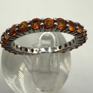 May include: A silver ring with a full band of round, orange gemstones. The ring is set on a clear, rounded surface, showcasing the gemstones' color and the ring's design. The gemstones are evenly spaced around the band.
