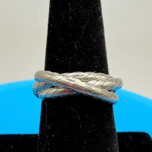 May include: A silver-colored ring with a braided rope design. The ring features two bands that cross over each other. The ring is displayed on a black velvet ring holder.